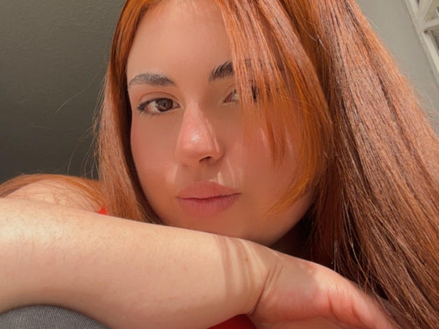 VicluX has orange hair, winking with a slight smirk, and rests her arm on a surface. Her light skin and subtle makeup are visible.