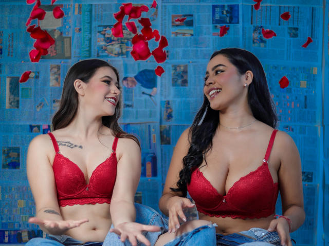 PaolaYKatty with big tits in red lace bras, laughing, sitting, wearing jeans, rose petals falling, smiling at each other, dark hair, tattoos, blue paper background, happy, relaxed.