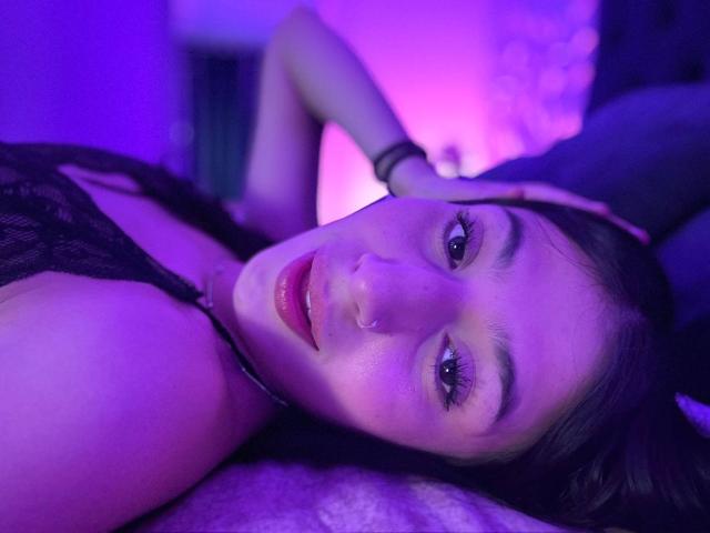 SophieBeaumont lying down with purple lighting, black lace top, hand in hair, looking at camera, subtle smile, pink lipstick, dark hair, soft skin, relaxed expression, cute, sexy.