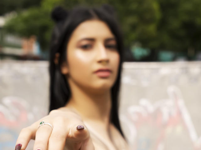 Blurry photo of AlisKooper with black hair in bear ears, pointing forward, showing a ring with a green gem and purple nail polish, soft-focused background.