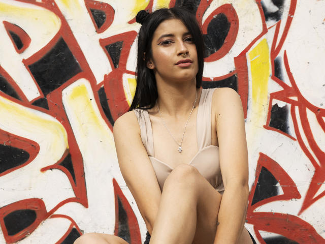 AlisKooper with dark hair, tan skin, and small tits, wearing a beige bra, sitting with legs crossed, looking at camera, wearing a necklace, graffiti background.