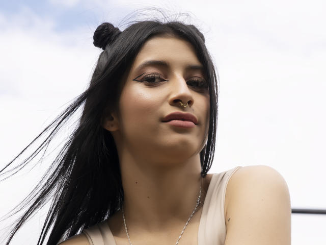 AlisKooper has black hair in a bun, nose ring, and winged eyeliner, wearing a beige top, with long hair blowing in the wind, under a cloudy sky.