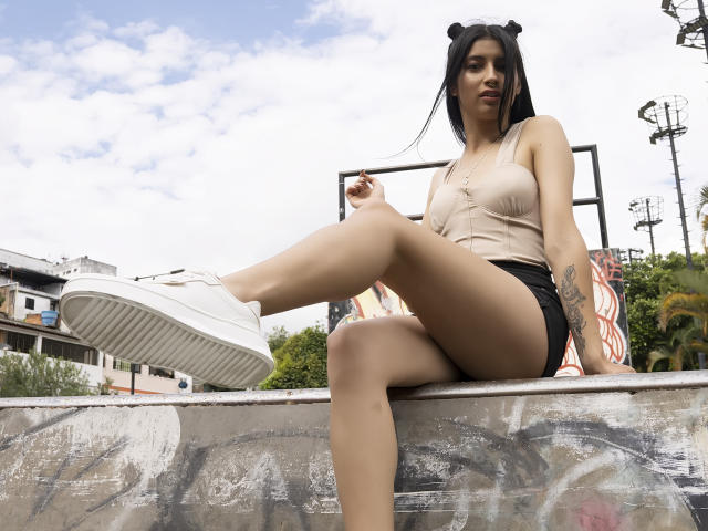 AlisKooper, with tan skin and black hair in bear ears, sits on a graffiti-covered wall. She's wearing a beige tank top, black shorts, and white sneakers. Her pussy is hidden.