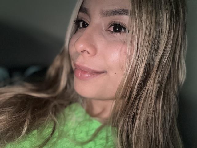AmaraRicci, met blond haar, een groene blouse en lichtjes open mond, lacht naar de camera. Haar ogen zijn donker en ze heeft een klein beauty markje op haar rechterwang..