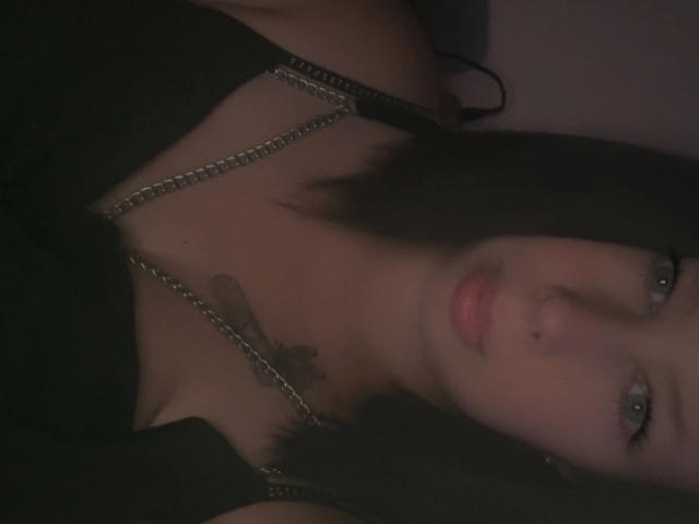 Siindyxx-hot with black hair, pale skin, and green eyes, wearing a black top with a metal chain, showing a tattoo on her chest, and slightly parted pink lips.