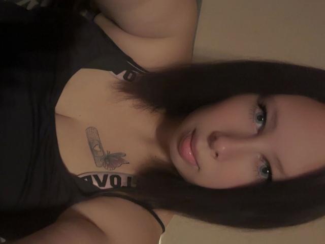 Siindyxx-hot with dark hair, green eyes, and a butterfly tattoo on her chest, wearing a black tank top, showing her tits, and a cute, slightly surprised expression.