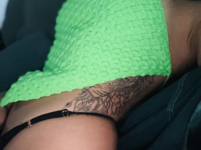 La schiena e i glutei di AmaraRicci, in una canotta verde a maglia, che mostra un tatuaggio nero dettagliato, indossa uno slip nero con dettagli dorati..