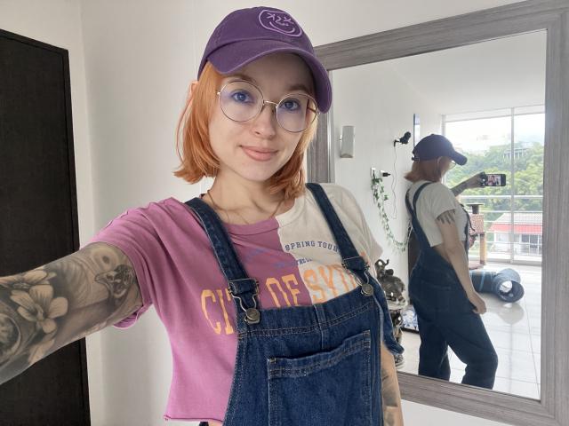 SarahRousen with orange hair, glasses, purple cap, purple shirt, and blue denim overalls, showing tattoos on her arm, smiling at the mirror, reflection of her back.