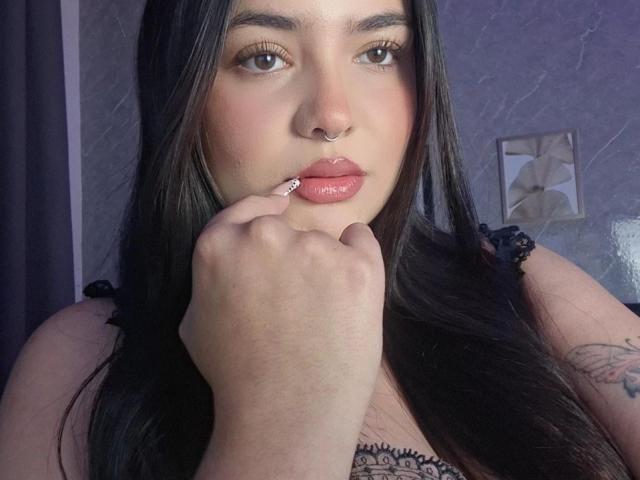 ValeriJoness has long black hair, a nose ring, and glossy pink lips. She's sucking on her finger with her hand near her face, showing a tattoo on her arm.