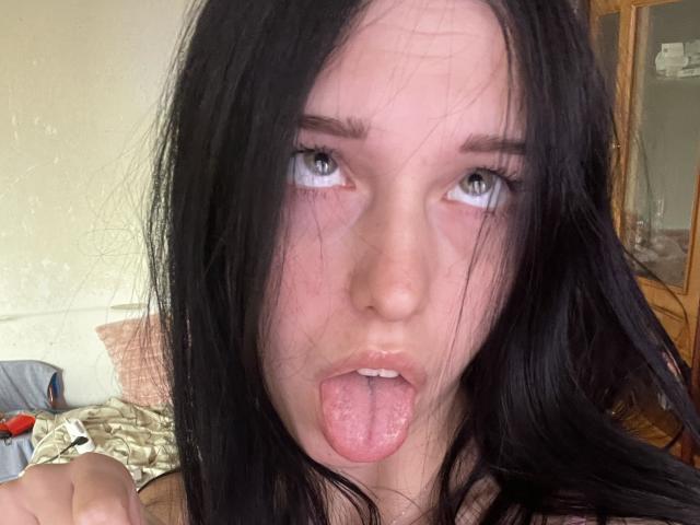 AmaiRin with black hair, blue eyes, and tongue out, showing her pink tongue and slightly parted lips, looking up with a slightly messy, tired face.