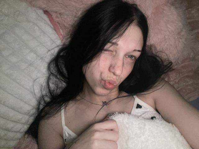 AmaiRin with long black hair, winking, pouting lips, holding a white stuffed toy, wearing a white tank top, lying on pink and white pillows.