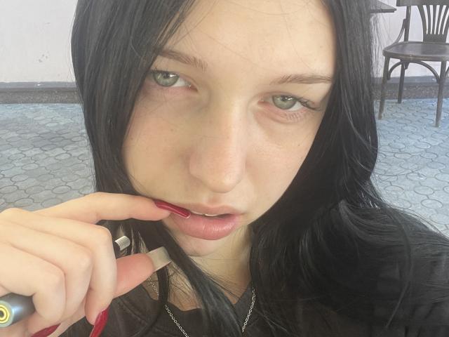 AmaiRin with black hair, green eyes, and red lipstick, holding a cigarette near her lips with a red-manicured finger, wearing a black top.