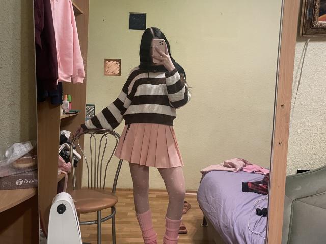 AmaiRin with long black hair, wearing a brown and white striped sweater, pink pleated skirt, pink socks, and no pants, taking a mirror selfie showing her ass.