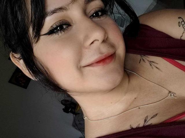 CandyI with dark hair, light skin, and subtle makeup, smiling, wearing a red top, necklace, and tattoos on her chest. Boobs visible under top, pink bra peeking.