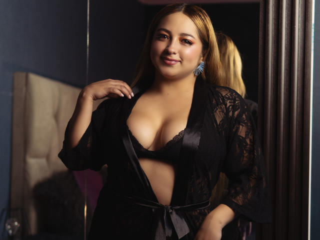 MaddyScarlett with blonde hair, wearing a black lace robe, exposing her tits in a low-cut black bra, smiling, hand on chest, blue earrings, in a dimly lit room.