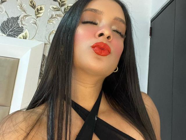 AileenDupont with long black hair, eyes closed, and red lipstick, puckering her lips, wearing a black criss-cross top, showing her tits, and a small gold earring.