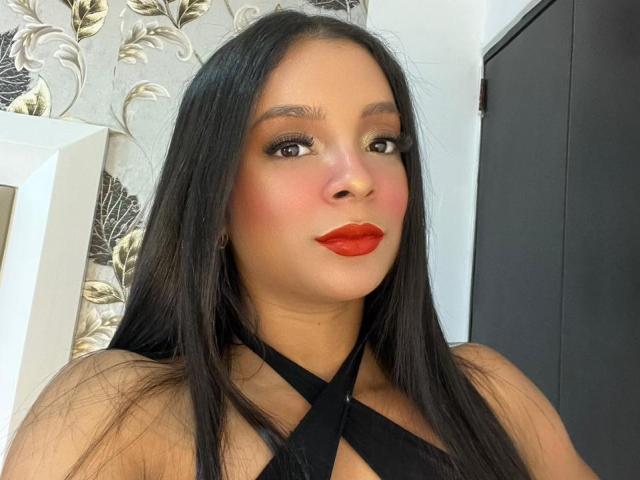 AileenDupont with long black hair, red lipstick, and black crisscross top, showing off her tits and a cute face, standing in front of a patterned wall.
