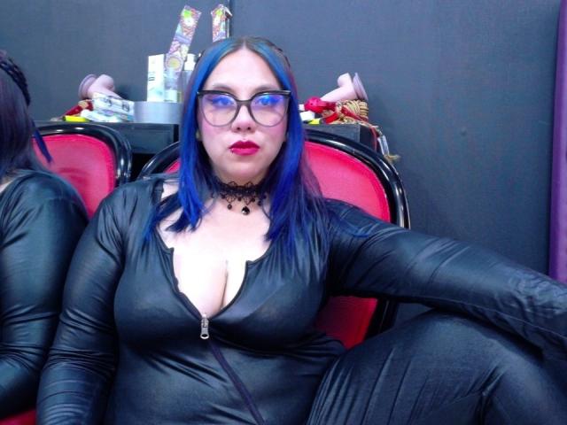 VelvetDomme with blue hair, glasses, red lipstick, and black leather zip-up outfit, showing her tits, sitting in red chair, arms resting on black armrests.