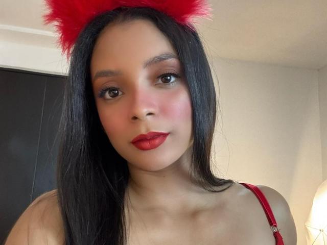 AileenDupont with red furry cat ears, red lipstick, and straight black hair, wearing a red strap dress, has a cute face and small tits. She looks at the camera.
