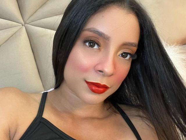 AileenDupont has long black hair, brown eyes, red lipstick, and a slight blush on her cheeks. She's wearing a black top, sitting on a beige, tufted couch.