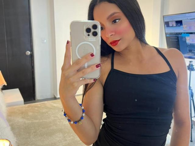 AileenDupont with long black hair, red lipstick, and black tank top, holding a phone with a ring grip, showing her red-painted nails and blue bead bracelet.