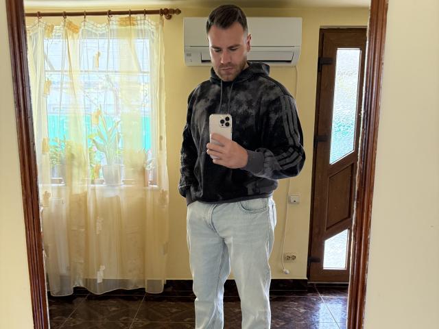 Thori69 stands in front of a mirror, taking a selfie. Thori69 has short brown hair, a beard, a black tie-dye hoodie, and light blue jeans. Thori69 holds a white iPhone.