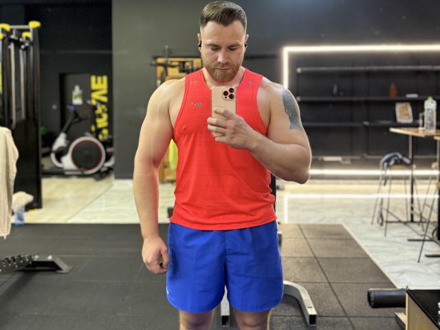Thori69, ein muskulöser Mann mit einem roten Tanktop und blauen Shorts, macht ein Selfie im Fitnessstudio. Er hat einen Bart, ein Tattoo auf seinem rechten Arm und ein ernstes Gesicht..