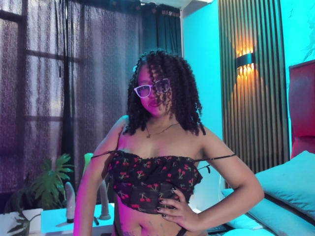 ArtemisaS with curly black hair, glasses, and a black cherry-patterned top, showing her tits and hand on hip, in a room with blue and orange lights.
