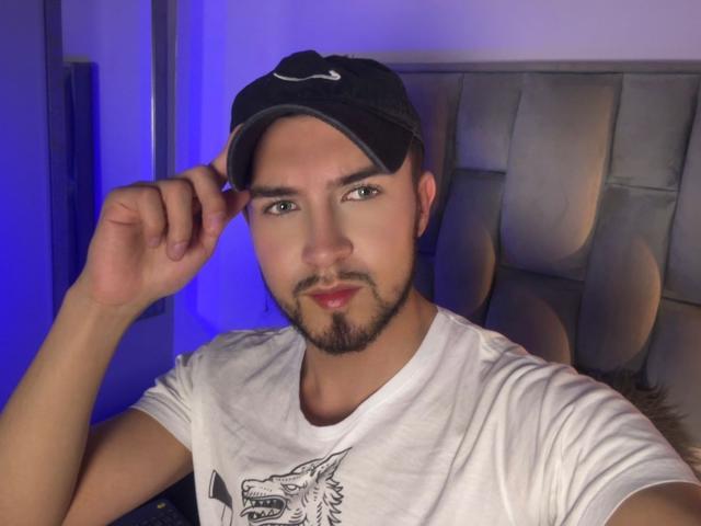DaltonWhite, che indossa un berretto nero, una camicia bianca con una stampa di lupi e una barba, tiene il berretto con la mano destra, con una luce blu sullo sfondo, ha un'espressione seria, è di corporatura media ed è attraente..