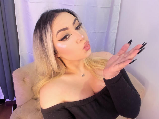 MelissaLust met blond haar, een zwarte top met een open schouder, lange nagels, volle lippen en een kleine tatoeage op haar borst, die haar hand omhoog steekt. Haar borsten zijn licht zichtbaar..