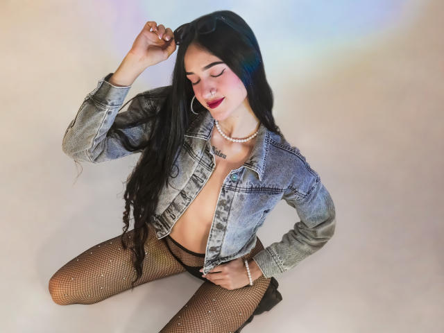 TaylorDiamons, with long black hair, wearing a denim jacket, pearl necklace, fishnet stockings, and black high heels, has her eyes closed, touching her hair, showing her tits.