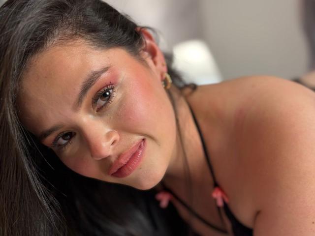 ValeriaNoir has dark brown hair, pinkish eyes, and a cute smile with pink lipstick. She's wearing a black necklace with pink bows, leaning forward. Her tits are partially visible.