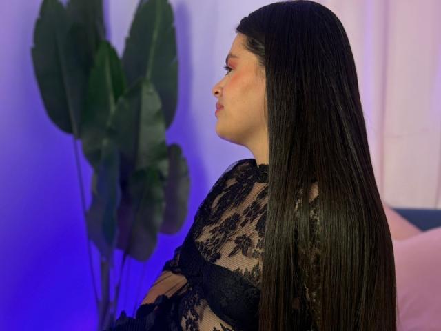ValeriaNoir with long black hair, wearing a black lace top, side profile, pouty lips, blue-purple light, green leafy plant in background. Her ass is partially visible.