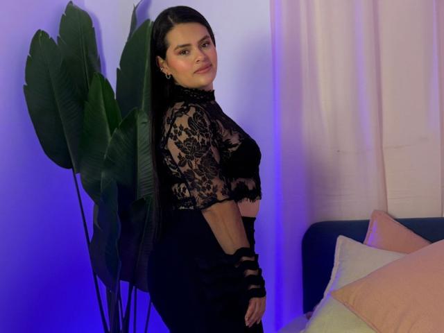 ValeriaNoir with long black hair, wearing a black lace top and black skirt, standing next to a green plant, showing her ass and a slight smile, looking back.