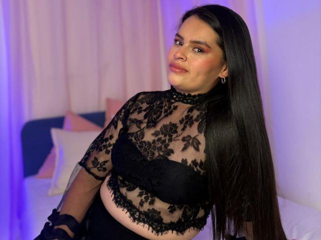 ValeriaNoir with long black hair, wearing a sheer black lace top showing her small tits and black bra, smiling slightly, in a bedroom with purple lighting.