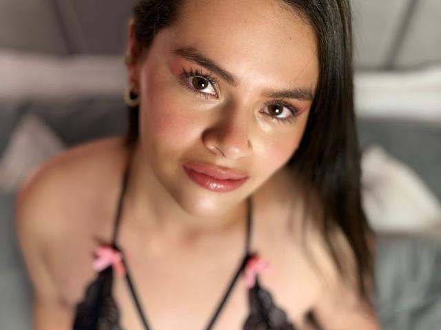 ValeriaNoir with glossy brown eyes, pink blush, and shiny lips, wearing black lace lingerie with pink bows, has small earrings, and long dark hair, looking up at the camera.