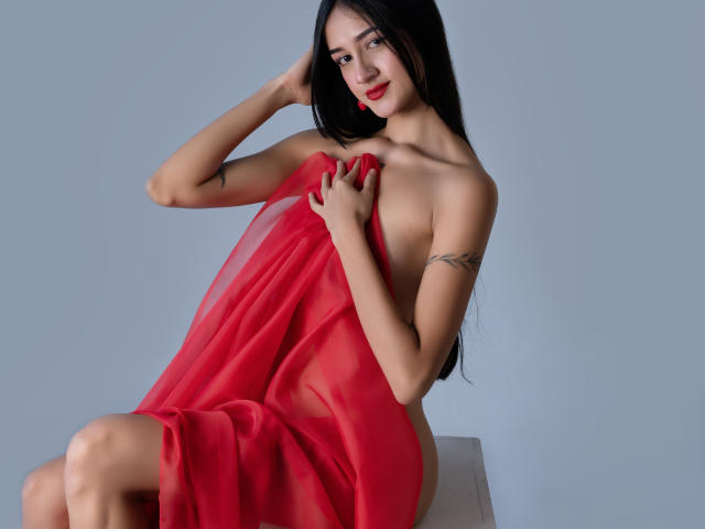 TaylorDiamons with long black hair, red lipstick, and tattoos, sitting on a chair, covering her tits with a red sheer cloth, smiling, hands touching her hair.