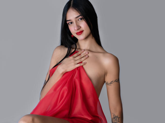 TaylorDiamons with long black hair, red lipstick, and tattoos, covering her tits with a red sheer cloth, smiling, hand on chest, gray background, sitting, looking at camera.