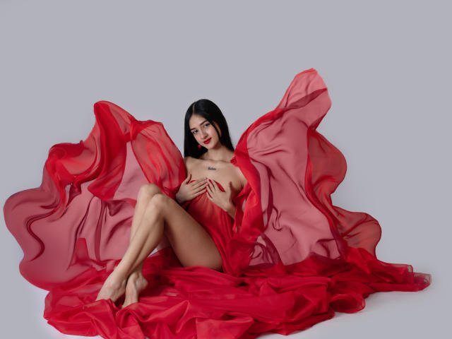 TaylorDiamons with red sheer fabric, covering her tits with one hand, sitting with legs crossed, bare-ass visible, looking at camera, long black hair, red lipstick, gray background.