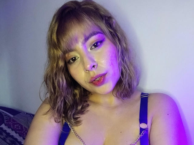 LolaEvans with wavy blonde hair, pink eyeliner, and purple light, wearing a blue top with gold chain straps, showing her tits and a nose ring.