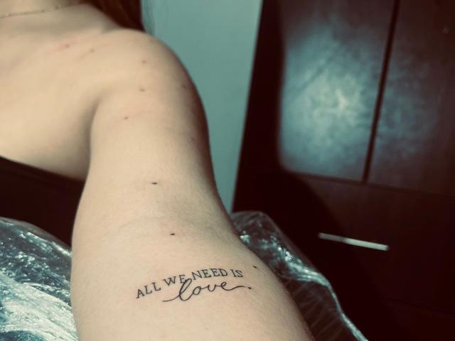 Il tatuaggio sulla parte superiore del braccio di OlivaStellas recita "TUTTO QUELLO DI CUI ABBIAMO BISOGNO È amore". La sua pelle chiara mostra piccole macchie e un leggero segno rosso vicino alla spalla..