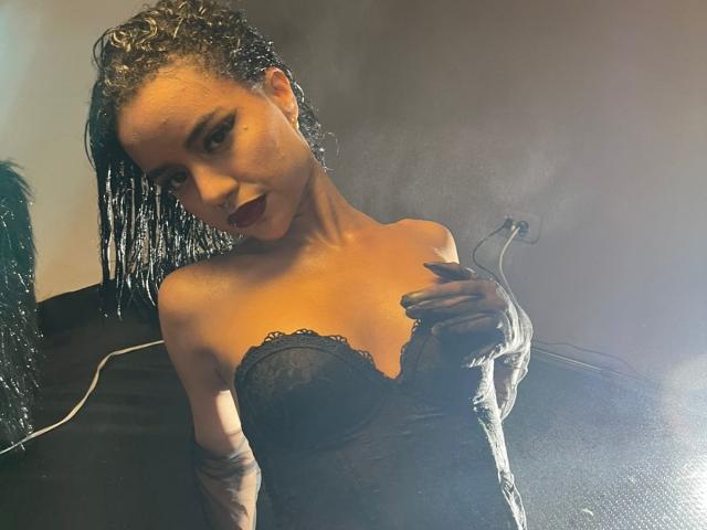 Wendy James, avec ses cheveux bouclés mouillés, un haut en dentelle noir sans bretelles, tenant une cigarette, regardant de manière séduisante, mettant en valeur ses seins et son bras tatoué, entourée de fumée..