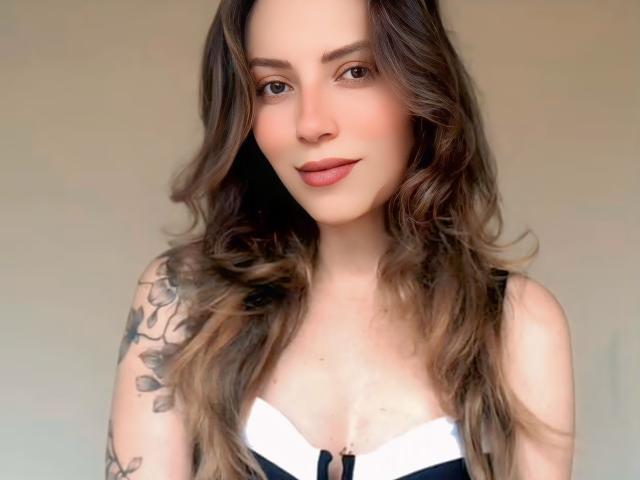 Belarara with wavy brown hair, light skin, and subtle makeup, wearing a black and white top, showing small tits and a tattoo on her right arm, smiling softly.