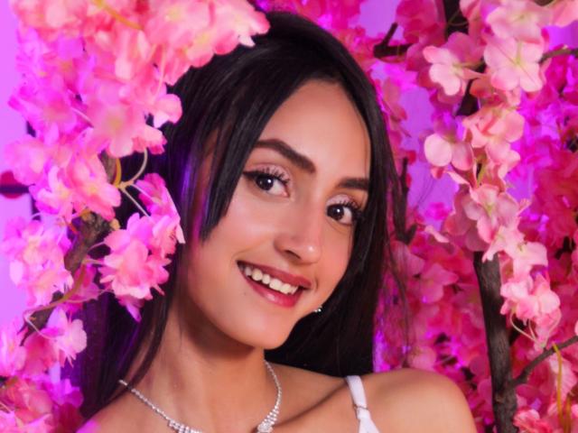 Meganoa with dark hair, smiling, pink cherry blossoms around, wearing white strap dress, silver necklace, purple highlight, cute, pretty, boobs partially visible, pinkish-purple background.