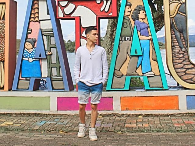 Santhiago69, staand, gekleed in een wit shirt met lange mouwen, blauwe en witte denim shorts en witte sneakers, voor een kleurrijke, grote muur met graffiti-achtige letters op een bakstenen muur..