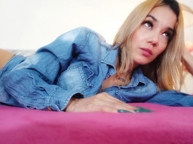 Veroo69 with blonde hair, nose ring, and green nail polish, lying on a pink bed, wearing a blue denim jacket, looking to the side with a slight pout.