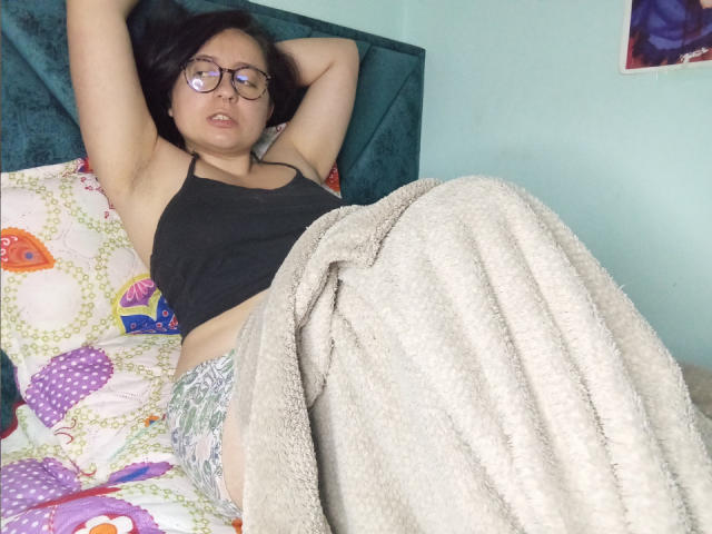 CristinaSmit, con gafas, camiseta negra y pantalones grises estampados, con los brazos por encima de la cabeza, acostada en la cama, una manta blanca y esponjosa cubriendo sus glúteos, cabecero verde, expresión relajada..