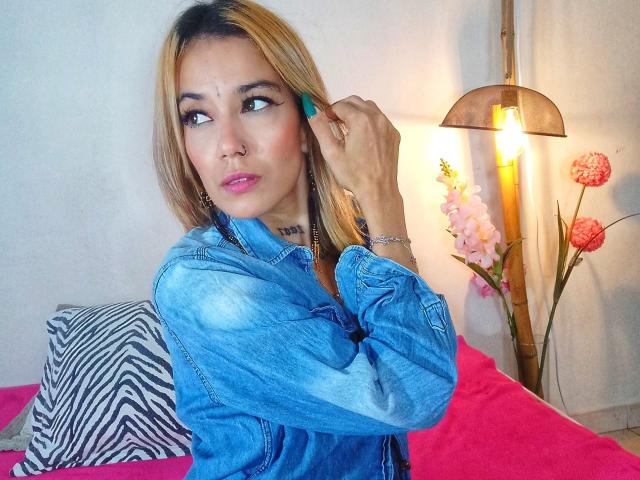 Veroo69 with blonde hair, blue denim jacket, pink lipstick, nose ring, green fingernail, zebra pillow, pink bed, bamboo lamp, and pink flowers in a cozy room.