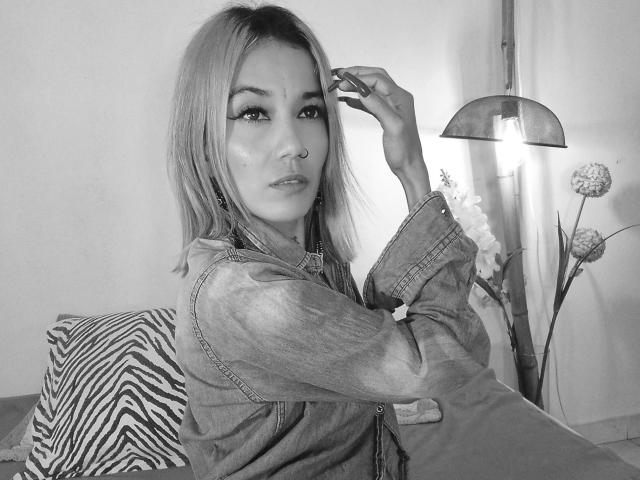 Veroo69 with blonde hair, wearing a denim jacket, nose ring, and eyeliner, touching her head, sitting on a zebra-patterned pillow, next to a lamp with a flower.