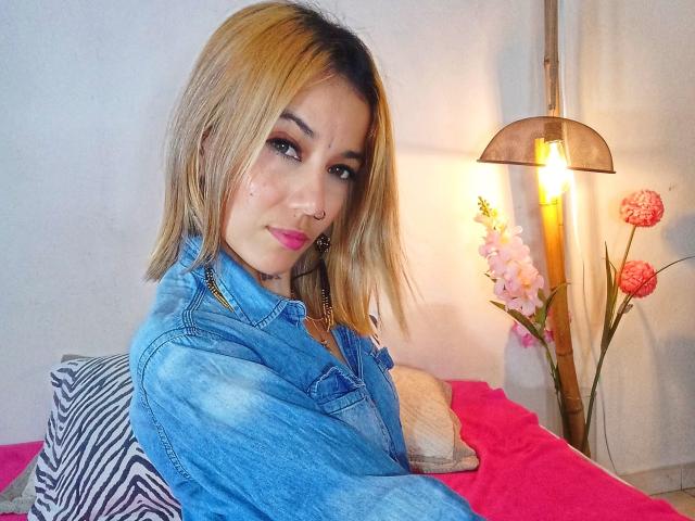 Veroo69 with blonde hair, denim jacket, pink lipstick, nose ring, and earrings, sitting on pink bed, lamp with pink flowers, and striped pillow in background.